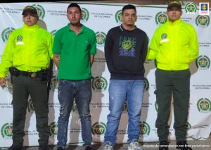 En la fotografía aparecen dos hombres capturados, acompañados de dos uniformados de la Policía. En la parte posterior un banner con logos de la entidad.