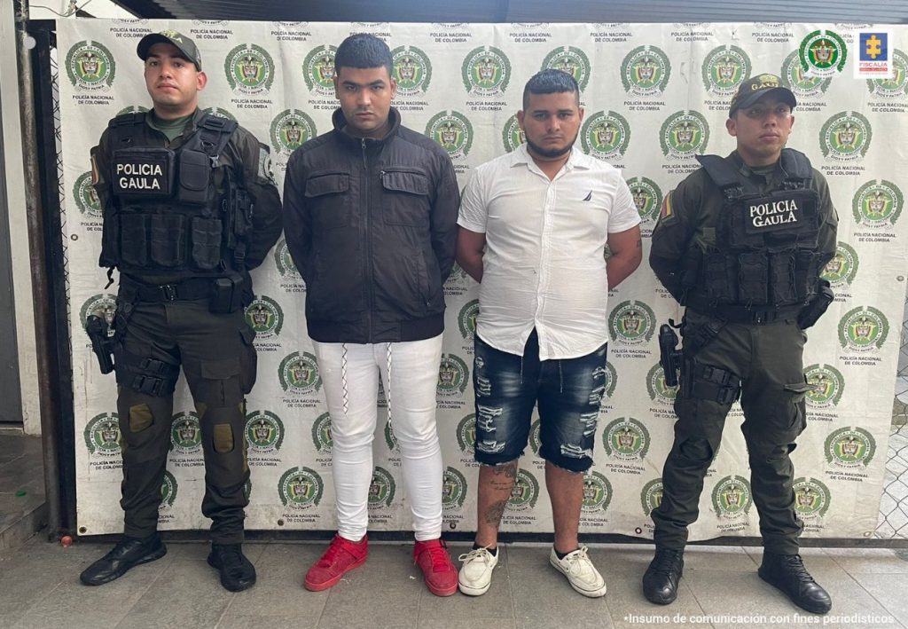 capturados de frente y esposados manos atrás, custodiados por dos uniformados de la Policía. Detrás banner de la Policía Nacional.