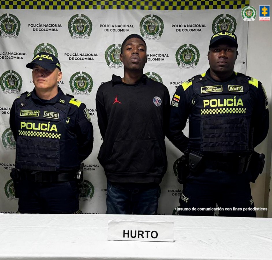  Se observa el capturado , de rente, a cada costado un funcionario de Policía Nacional, atrás , un pendón de Policía Nacional, adelante, una mesa con el cartel HURTO