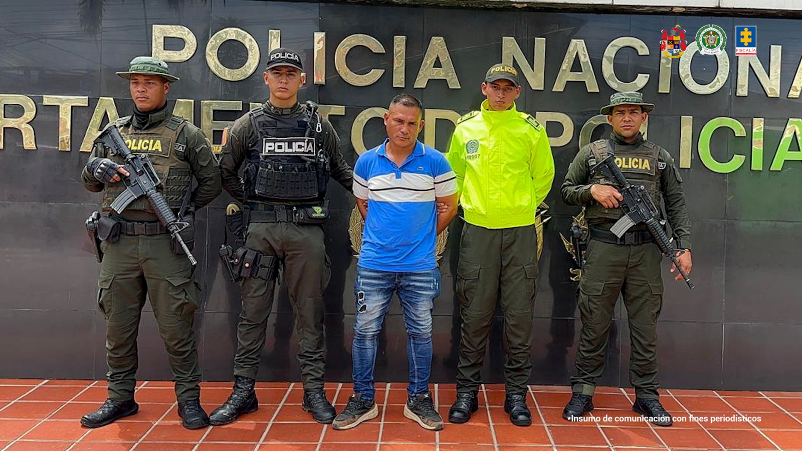 El capturado aparece de pie, con las manos esposadas en la espalda. Está rodeado por cuatro miembros de la Policía. Al fondo hay un letrero que dice Policía Nacional.