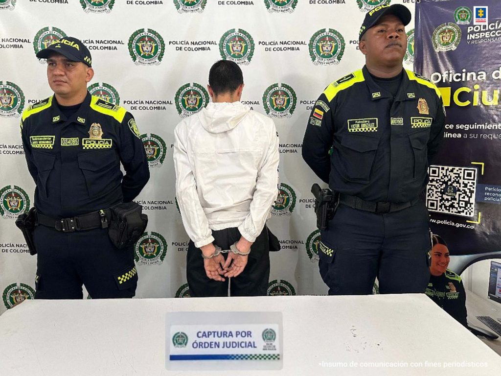 En la fotografía se aprecia de pie, con los brazos atrás, esposado y de espaldas a la cámara a Duván Sebastián Rodas Narváez. Viste buso blando de manga larga, con capucha y pantalón negro. A lado  y lado de él hay uniformados de la Policía Nacional. En la parte frontal hay una superficie blanca y sobre ella un letrero con logos de la Policía que dice: ‘Captura por orden judicial’. En la parte posterior, hay pendones institucionales de la Policía.