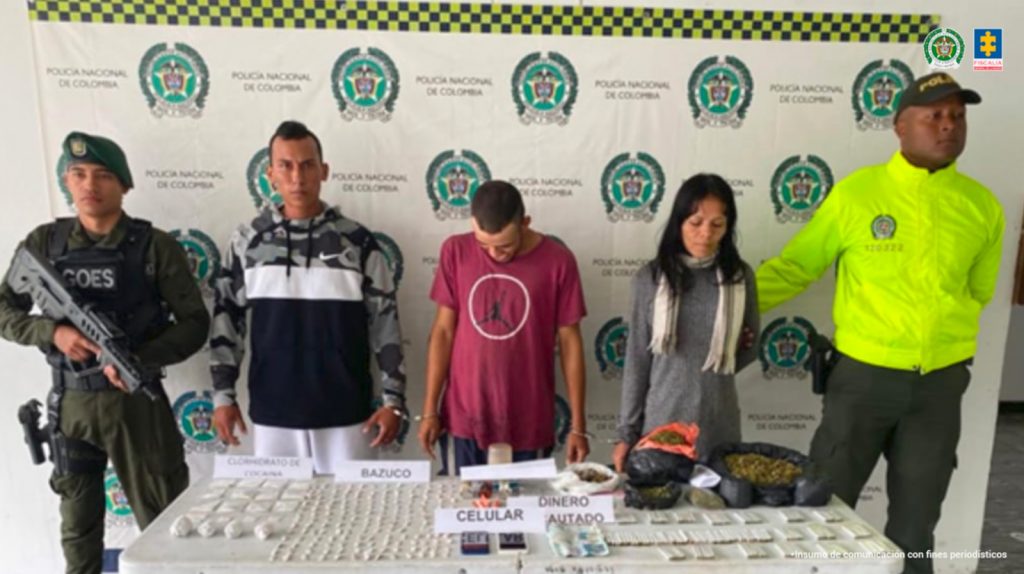 En la fotografía se aprecian de pie esposados entre sí a Duvian Alexis Rojas Cuartas; Duberney Cobo Izquierdo y Nazly Elizabeth Rengifo Cárdenas. La mujer viste blusa de color gris, de manga larga; con bufanda de color beage en el cuello y usa cabello largo. Otro, viste de camiseta de color vinotinto, de manga corta con cículo blanco estampado y una silueta negra de un hombre saltando; así como pantalón azul oscuro. El otro hombre viste buso de color negro con mangas con estampado camuflafo de color gris y negro; pantalón blanco. A los extremos de ellos hay uniformados de la Policía Nacional. Delante de ellos hay una superficie de color verde y sobre ella hay bolsas transparentes con contenido de color blanco y beage; así como bolsas negras con contenido vegetal; cigarrillos, dinero y celulares. Detrás hay un pendón institucional de la Policía.