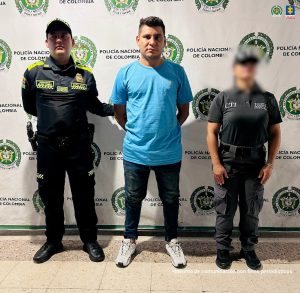 En la foto aparecen el capturado con buso azul de frente y custodiado por un servidor del Cuerpo Técnico de Investigación CTI y la Policía Nacional. En la foto aparece un backing de la Policía Nacional.