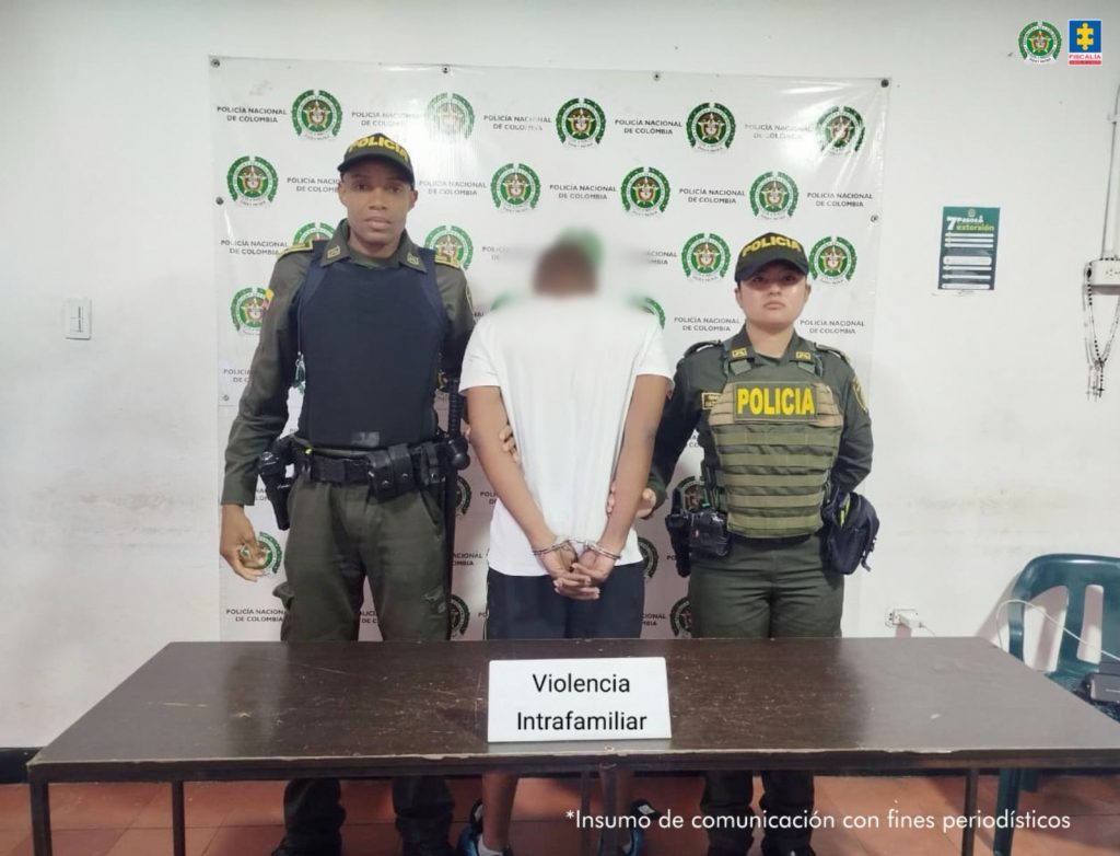 En la fotografía aparece de espaldas a la cámara, pie, con los brazos atrás y esposado el hoy imputado. Viste con camiseta de manga corta, de color blanco y pantalón negro. A sus extremos uniformados de la Policía Nacional. Delante, hay una mesa de color marrón y sobre ella hay un letrero que dice: ‘Violencia intrafamiliar’. Detrás de ellos hay un pendón institucional de la Policía.