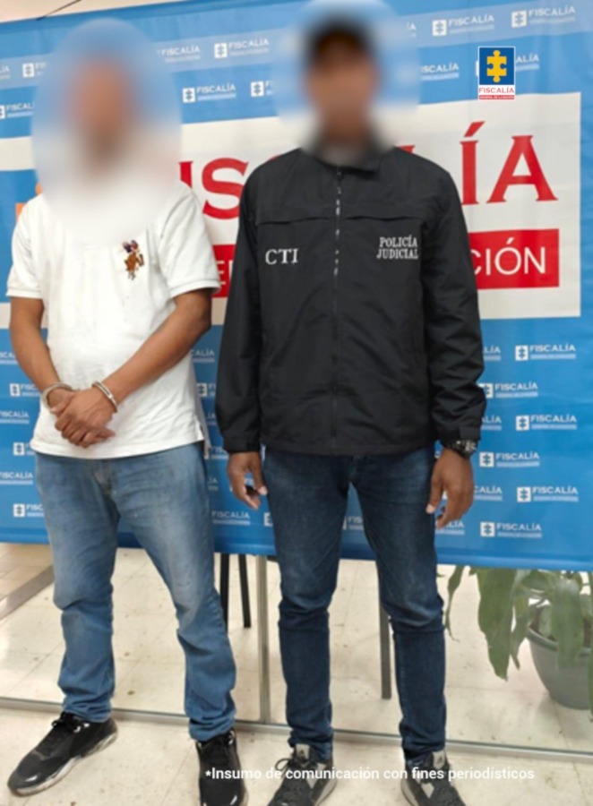 En la fotografía aparece el presunto implicado de pie, con los brazos adelante y esposado. Viste con camisa blanca, de manga corta, jean azul y zapatillas negras con blanco. A su extremo izquierdo hay un servidor del CTI de la Fiscalía. Detrás de ellos hay un pendón institucional de la Fiscalía.