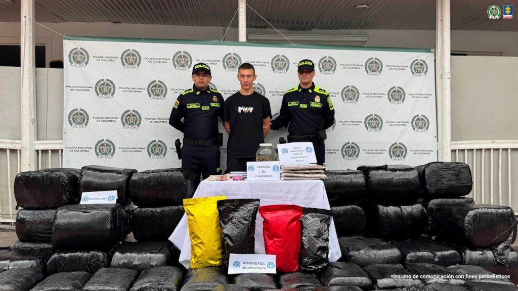 Se observa al capturado en el centro y a cada costado un funcionario de Policía Nacional que lo custodian. Al frente, una mesa . Abajo se observan varias bolsas negras y de colores que contiene el estupefaciente.