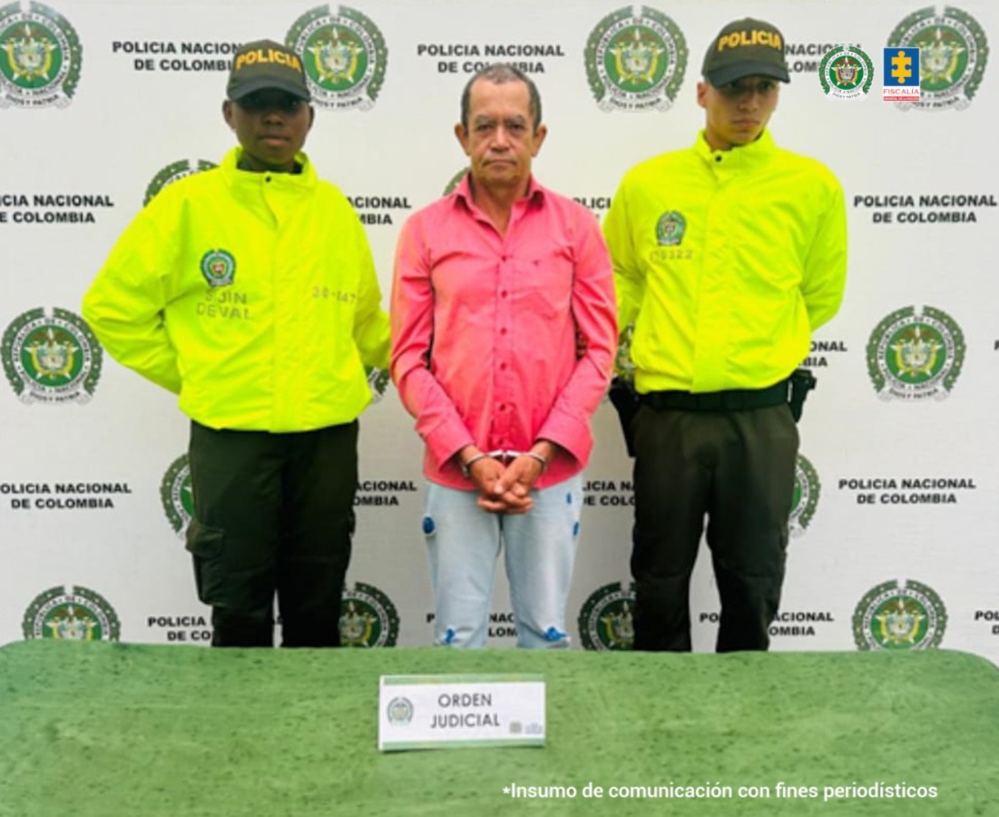 En la fotografía se aprecia de pie, con los brazos adelante y esposado a Edelberto Jaramillo Acuesta. Viste camisa de manga larga de color rosado y jean azul claro desgastado. A sus extremos hay uniformados de la Policía Nacional. Delante de ellos hay una superficie de color verde y sobre ella hay un letrero que dice: ‘Orden Judicial’. Detrás de ellos hay un pendón institucional de la Policía.