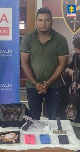 En la imagen aparece una persona de suéter verde capturada frente a una mesa donde hay una pistola, tres celulares y un cuaderno.