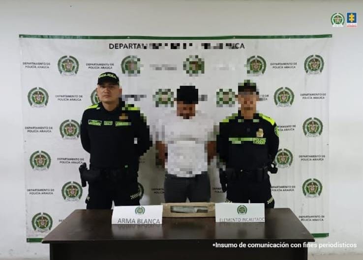 Se visualiza al capturado junto a dos uniformados de la Policía Nacional. Frente a ellos una mesa con el arma incautada. Detrás de ellos el banner que identifica a la Policía Nacional.