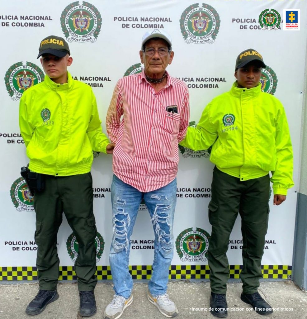 Está el capturado de frentes con gorra, esposado manos atrás, custodiado por dos policías uniformados. Detrás banner de la Policía Nacional.