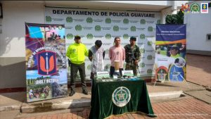 capturados de frente custodiados por un policía y un militar, delante de ellos una mesa con material incautado. Detrás banner de la Policía y del Ejército Nacional.