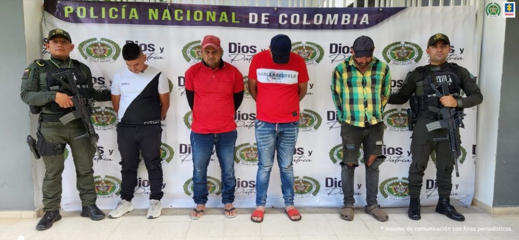 capturados de frente, esposados manos atrás custodiados por dos policías. Detrás banner de la Policía Nacional.