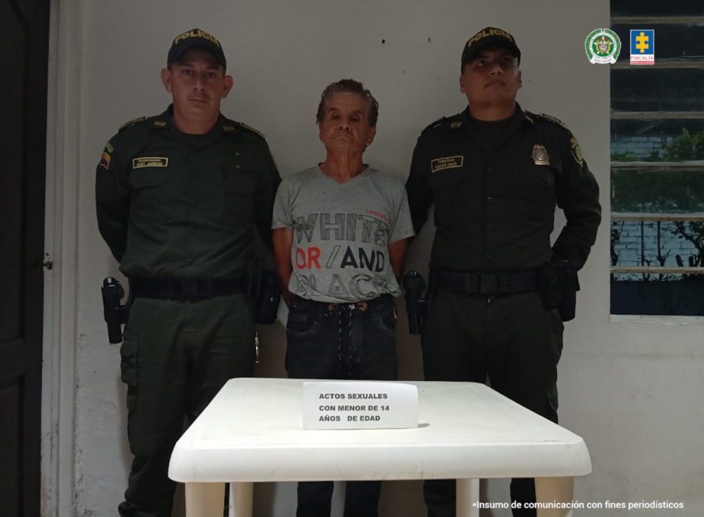En la fotografía se aprecia de pie, con los brazos atrás Edilberto Ríos Bernal. Viste camiseta de manga corta de color gris con letras estampdas en rojo, hegro y gris; pantalón oscuro. A sus extremos hay uniformados de la Policía Nacional. Delante de ellos hay una pesa blanca y sobre ella un letrero que dice: ‘Actos sexuales con menor de 14 años de edad’.