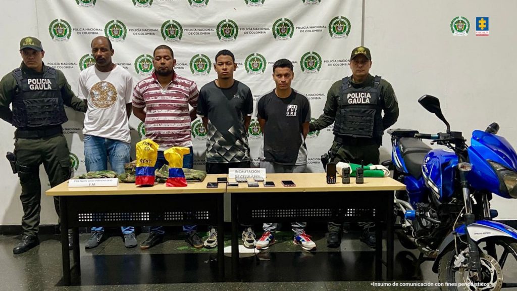 En la imagen están los 4 procesados con dos servidores del Gaula de la Policía a los lados y una mesa adelante con celulares, radios de comunicación, uniformes camuflados del Ejército Nacional y brazaletes de la bandera de Colombia con mensajes alusivos a las Farc y al lado una motocicleta azul, todos estos elementos fueron incautados en las diligencias de allanamiento.