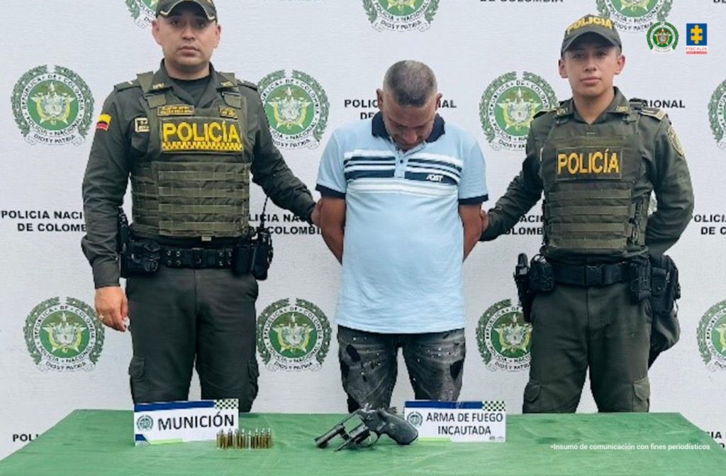 En la fotografía aparece de pie, con los brazos atrás y la cabeza inclinada hacia adelante. Viste con de color azul claro, cuello d color azul oscuro, frajas azules en los bordes de las mangas y franjas azules y blancas en el pecho; jean de color gris desgastado. En sus extremos hay uniformados de la Policía Nacional. Delante de ellos hay una superficie de color verde y sobre ella hay un arma de fuego y munición. Asimimso, hay dos letreros que dicen: ‘Munición’ y ´Arma de fuego incautada’. En la parte de atrás hay un pendón institucional de la Policía.