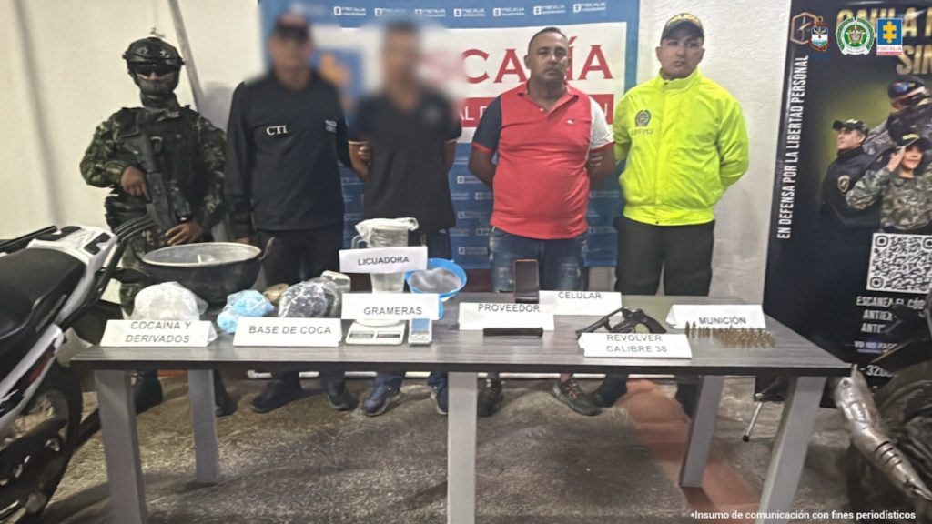 En la imagen aparece una persona de suéter rojo, entre uniformados del CTI, la Policía y el Ejército Nacional frente a una mesa donde hay munición, un revólver, una licuadora, una gramera y estupefacientes.