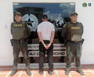 capturado de espaldas esposado, custodiado por dos uniformados de la Policía Nacional. Detrás banner de la Policía Nacional.
