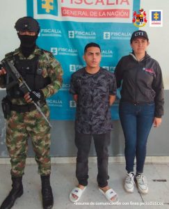 capturado de frente manos atrás custodiado por Investigadora del CTI de la Fiscalía y Militar. Banner de Fiscalía.