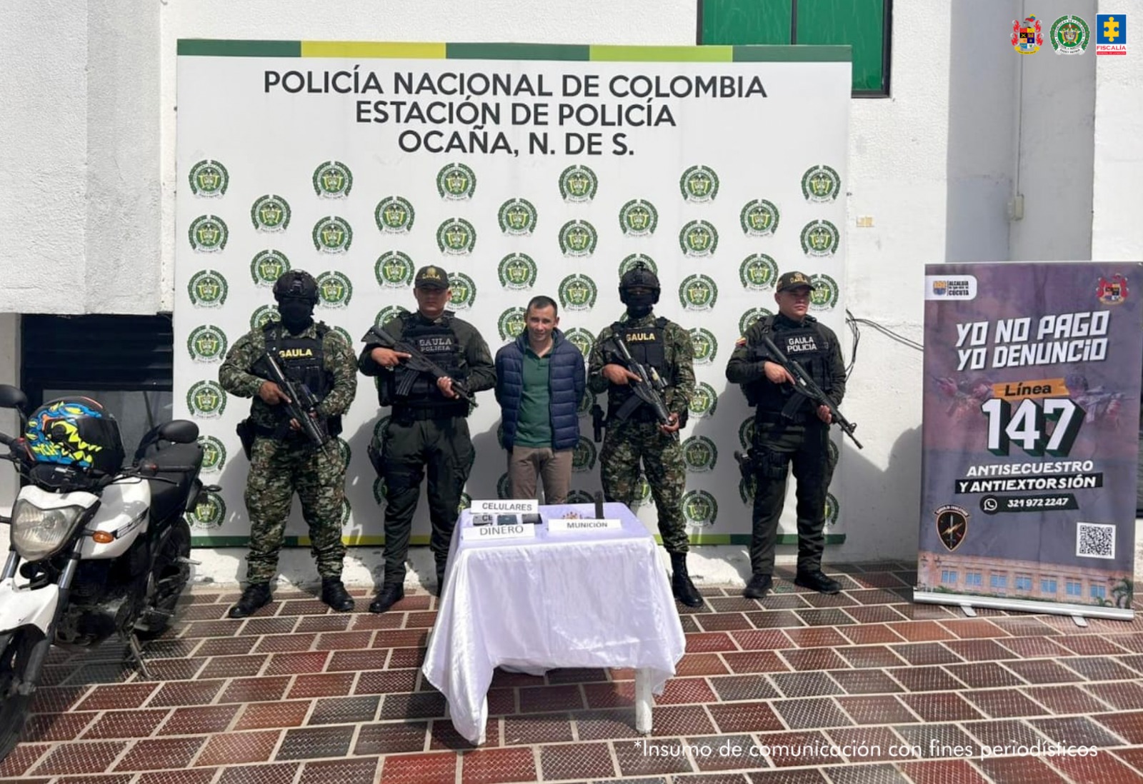 En la foto se ven cinco personas, una de ellas el capturado y cuatro uniformados de la Policía y el Ejército Nacional. En la parte posterior se ve un backing de la Policía y en el lado derecho otro backing con una línea de denuncia de extorsiones del Ejército. 