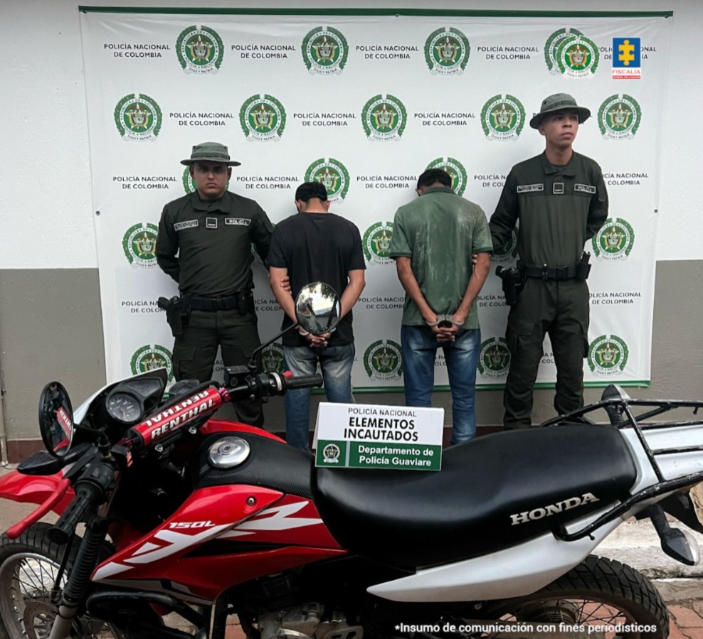 capturados de espaldas custodiados por dos policías, delante la motocicleta incautada. Detrás banner de la Policía Nacional.