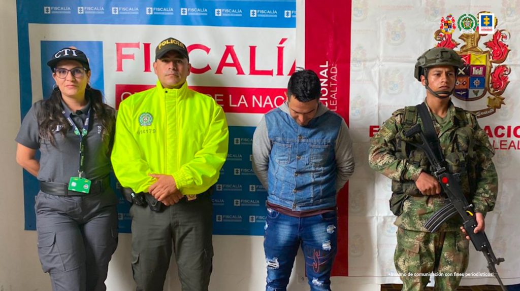 En la fotografía aparece un capturado junto a personal de Fiscalía General de La Nación, Ejército Nacional, Policía Nacional. En la parte posterior de la imagen se ven banners de Ejército Nacional, Policía Nacional y la Fiscalía General de la Nación.