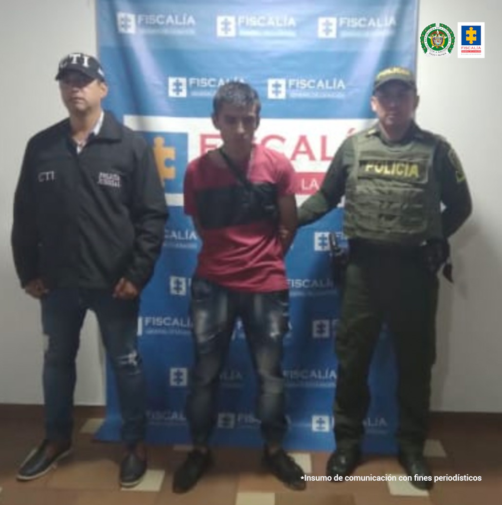 En la foto aparece el judicializado con las manos atrás, vestido con camiseta roja, jean azul y zapatos negros. Al lado derecho del judicializado aparece un servidor del CTI de la Fiscalía y al lado izquierdo de él aparece un integrante de la Policía Nacional. Detrás de ellos aparece un pendón institucional de la Fiscalía.