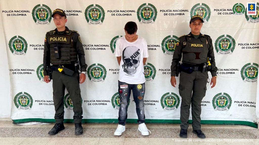 En la fotografía aparece un capturado, junto a personal de Policía Nacional. En la parte posterior de la imagen se ven banners de Policía Nacional y la Fiscalía General de la Nación. 