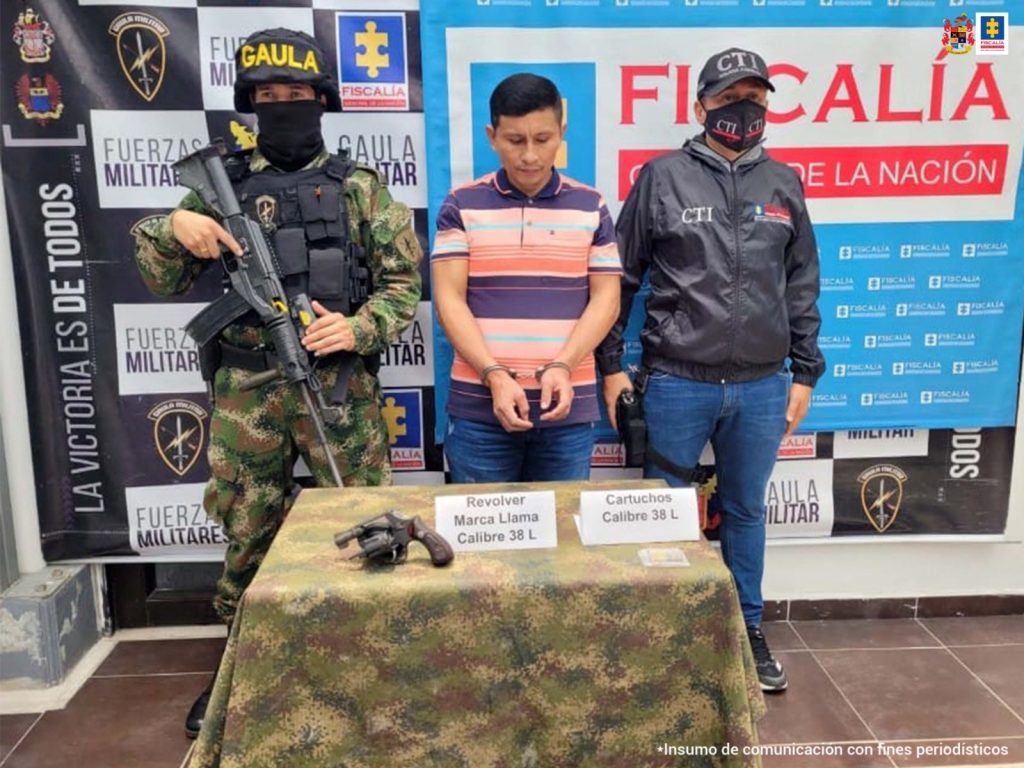 capturado esposado de frente custodiado por investigador del CTI de la Fiscalía y militar del Ejército Nacional. Delante de ellos una mesa con arma y munición incautaos al detenido, detrás banner de fiscalía y de Gaula Militar.
