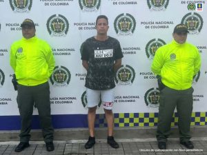 En la foto aparece, de pie y con los brazos atrás, Elio David Mogollón Medrano. Viste camiseta de manga corta verde estampada; bermuda azul clara y tenis negrosl. A lado y lado de ellos hay uniformados de la Policía Nacional. Detrás de ellos hay un pendón institucional de la Policía.