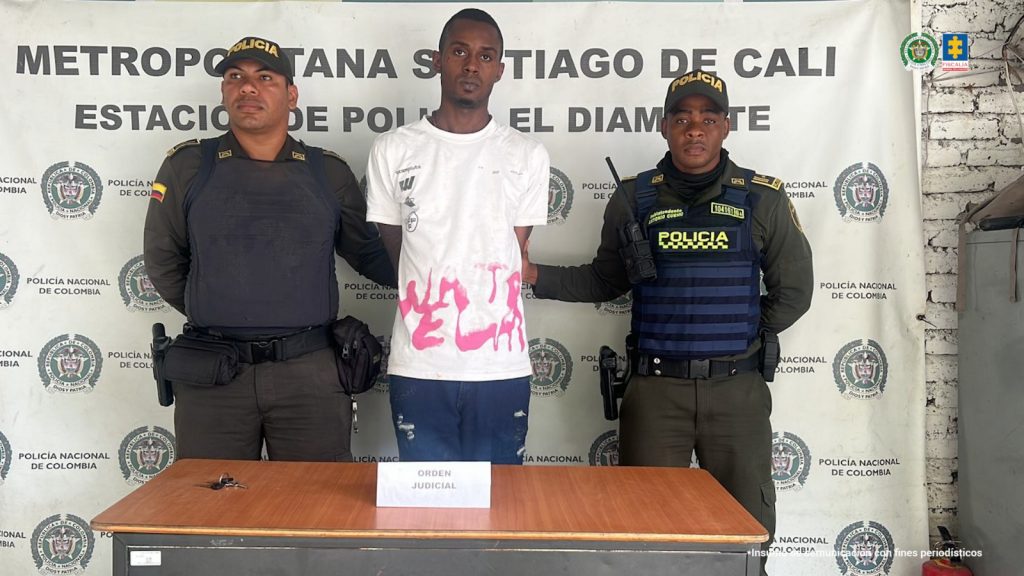 En la fotografía se aprecia de pie y con los brazos atrás a Elver Jesús Castillo Sinisterra, alias Yupi. Usa Chivera y viste con camiseta blanca, manga corta y con estampado fucsia en la parte inferior; así como jean azul. A sus extremos hay uniformados de la Policía Nacional. Delante de ellos hay una superficie de color marrón y sobre hay un letrero que dice: ‘Orden judicial’. Detrás de ellos hay un pendón institucional de la Policía.