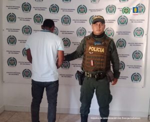 El imputado se encuentra de espaldas y tiene un buso blanco y jean, se encuentra en custodia por un uniformado de la Policía Nacional. Detrás se observa un backing de la Policía.