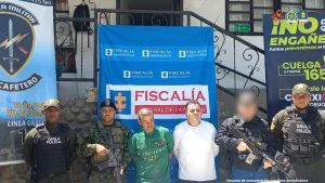 Se observan una fila con los dos capturados de frente, uno al lado del otro, uno viste camiseta verde y el otro camiseta blanca. A cada lado funcionarios del CTI, Gaula Policía y Gaula Militar. Atrás quedó un pendón de Fiscalía General de la Nación, uno de Gaula Policía y otro de Gaula Militar.