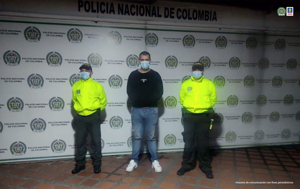 Se observa al detenido en el centro y a sus dos lados un funcionario de la Policía Nacional. En el fondo un pendón de la Policía Nacional
