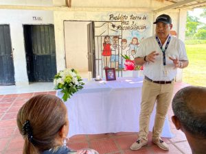En la imagen se ve un funcionario hablando, frente a una mesa y lo observan dos personas.