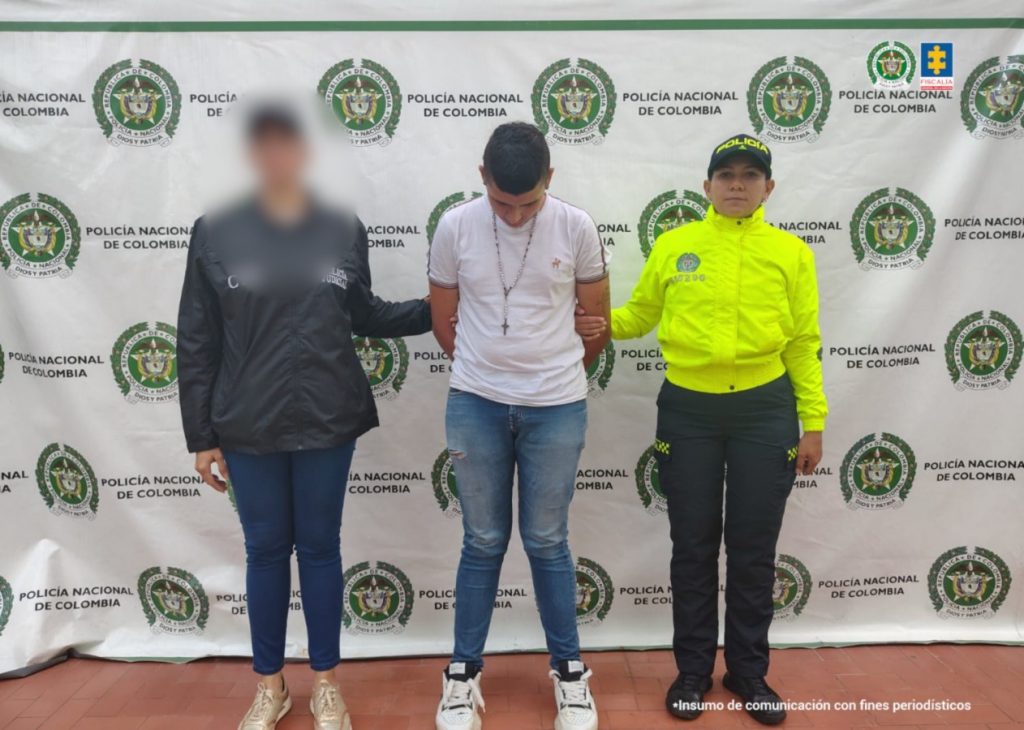En la foto se observa al capturado custodiado por una servidora del Cuerpo Técnico de Investigación CTI y una uniformada de la Policía Nacional. También se observa un backing de la Policía Nacional. El capturado tiene unos tenis blancos, una camándula y buzo blanco.