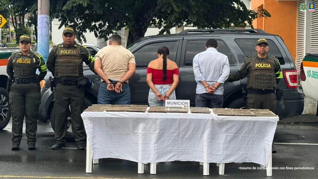 La fotografía fue tomada en exteriores y aparecen de espaldas a la cámara, con los brazos atrás y esposados: Erdin Jesús Saavedra Muñoz, Karen Daniela Enríquez Rincón y Carlos Eduardo Rodríguez Rodríguez. Uno, viste con camiseta de mangas cortas de color beage y jean azul; la mujer, usa cabello largo con trenza y viste con blusa de color rojo, con hombros destapados, mangas cortas y jean azul claro. La otra persona viste con camisa de mangas largas, de color azul claro y jean gris oscuro. En los extremos de ellos hay uniformados de la Policía Nacional. En la parte delantera hay una mesa con mantel blanco y sobre ella se aprecia gran cantidad de munición y un letrero que dice: ‘Munición’. En la parte posterior de la fotografía hay un vehículo de color azul oscuro.