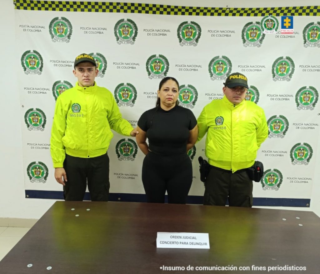 En la fotografía se aprecia de pie y con los brazos atrás a Erika Andrea Varela Pedroza, alias Erika. Viste camiseta y pantalón de color negro. A sus extremos hay uniformados de la Policía Nacional, delante de ellos hay una superficie de color marrón y sobre ella hay un letrero que dice: ‘Orden judicial Concierto para delinquir’. Detrás de ellos hay un pendón institucional de la Policía.