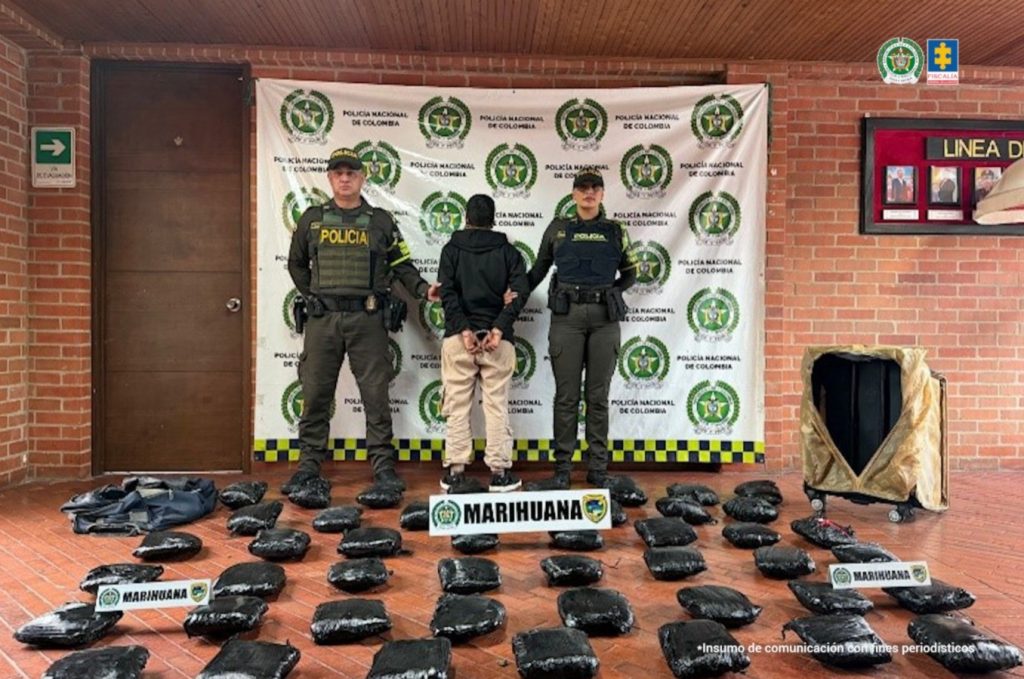 En la fotografía aparece de pie, de espaldas a la cámara, con los brazos atrás y esposada Erika Brillyth Acuña Castro. Viuste con buso negro y pantalón de color beage. Usa cabello corto. En sus extremos hay uniformados de la Policía nacional. Sobre el suelo hay varios paquetes envueltos en papel plástico negro, una maleta y dos letreros que dicen: ‘Marihuana’. En la parte posterior hay un pendón institucional de la Policía.