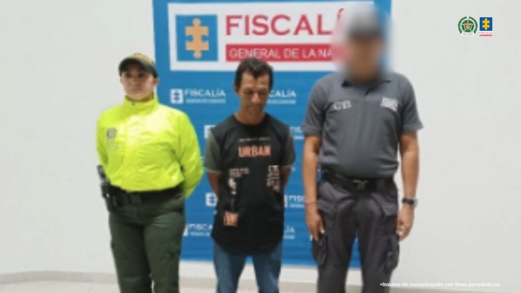 Se observa al capturado de frente, a un costado, un funcionario del CTI y al otro uno de Policía Nacional.  Atrás, un pendón de la Fiscalía General de la Nación.