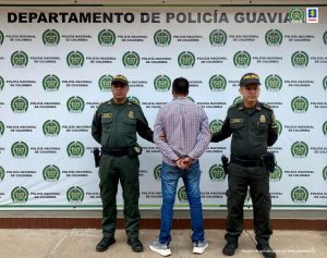 Capturado de espaldas esposado, custodiado por dos uniformados de la Policía Nacional. Detrás banner de la Policía.