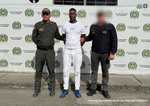 En la fotografía se aprecia de pie y con los brazos atrás a Esteban Riascos Hernández. Viste con camiseta de manga corta y pantalón de color blanco; zapatillas azules con negro. A su extremo izquierdo hay un servidor del CTI de la Fiscalía y al derecho un uniformado de la Policía Nacional. Detrás de ellos hay un pendón institucional de la Policía.