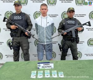 En la fotografía aparece de pie y con los brazos atrás Esteban Valencia Marulanda. Viste con buso de sudadera blanco con estampado gris en amndas y torso; pantalonea gris con estampado negro tipo cebra. Usa cabello rapado. En sus extremos hay uniformados del Gaula de la Policía Nacional. Delante de ellos hay una superfice verde y sobre ella se aprecia dinero en efectivo y un letrero que dice: ‘Dinero incautado’. En la parte posterior hay un pendón institucional de la Policía.