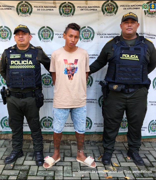 En la imagen aparece una persona de bermuda de jean y suéter beige, entre dos uniformados de la Policía Nacional.