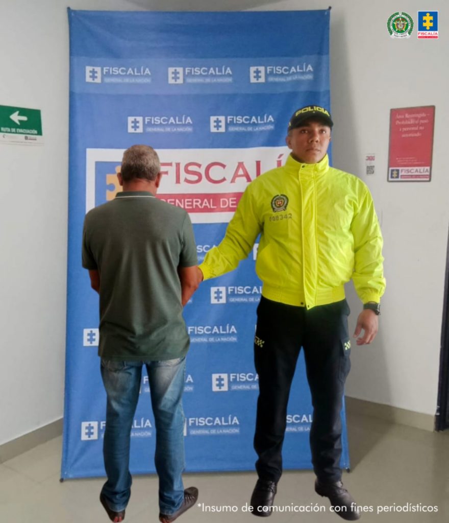 Se visualiza al capturado de espaldas junto a un uniformado de la Policía Nacional. Detrás de ellos el banner que identifica a la Fiscalía General de la Nación.