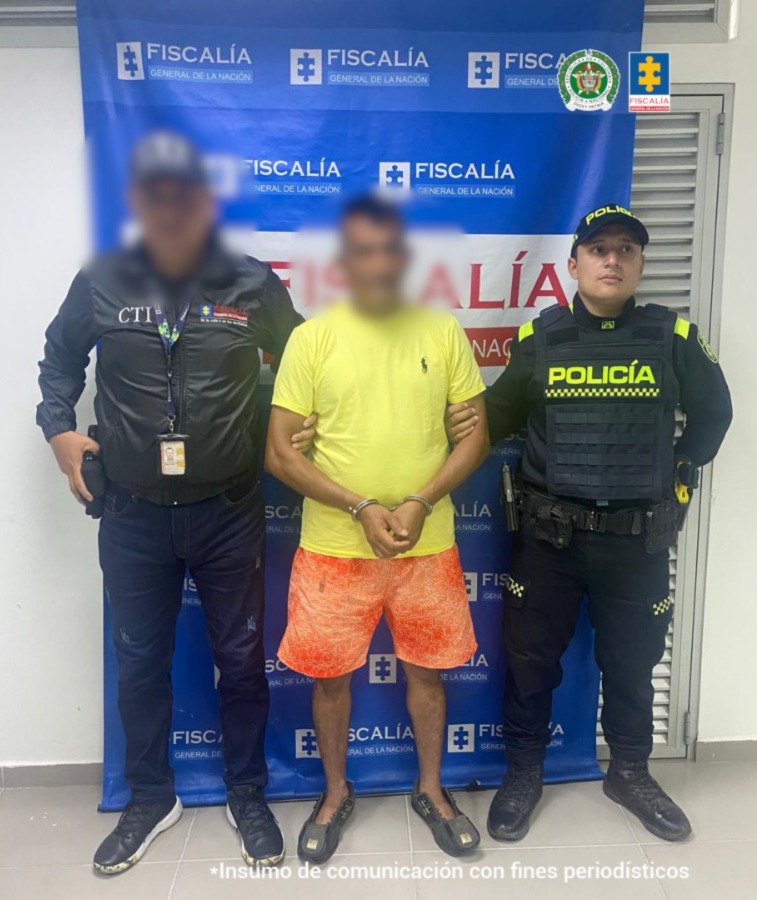 capturado de frente custodiado por investigador del CTI de Fiscalía y policía. Detrás banner de la Fiscalía.