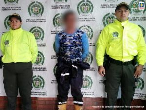 Capturado de frente custodiado por dos uniformados de la Policía. Detrás banner de la Policía.