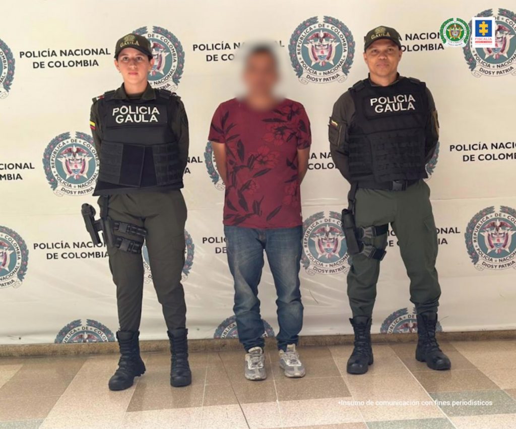 AL CENTRO SE OBSERVA UN CAPTURADO CON CAMISETA VINOTINTO. SOBRE EL ROSTRO UN EFECTO DE DIFUMINADO. A CADA COSTADO UNIFORMADOS DE POLICIA NACIONAL Y AL FONDO UN PENDÓN DE POLICÍA NACIONAL