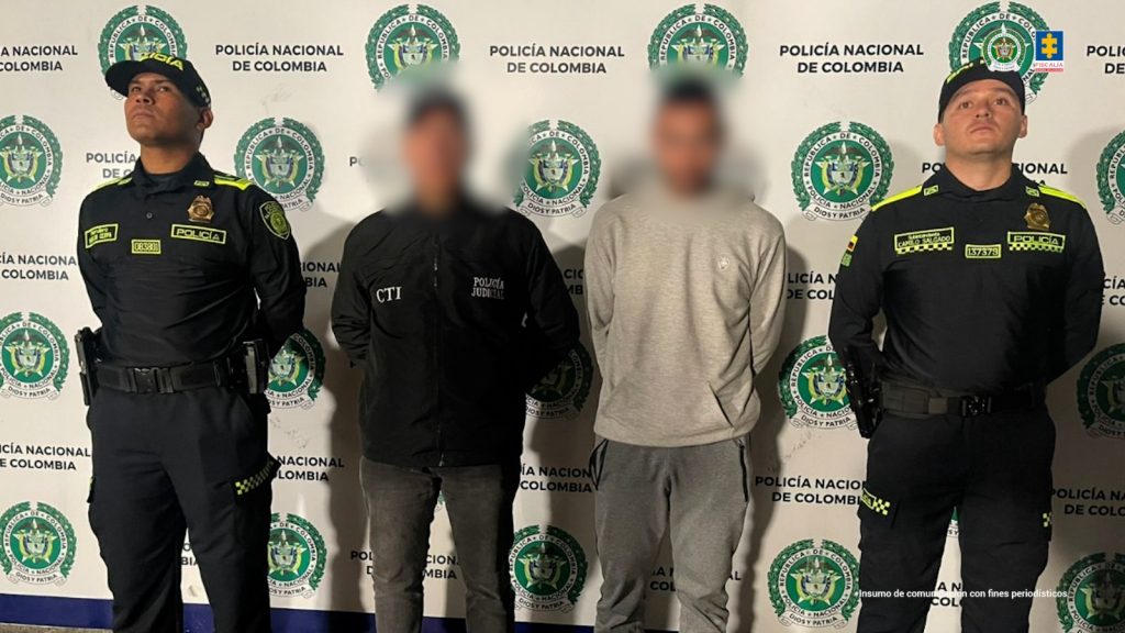 Se observa al capturado de frente, su rostro se ve difuminado debido a un efecto que lo hace borroso. A un costado lo custodia un funcionario del Cuerpo Técnico de Investigación CTI con uno de Policía Nacional. Al otro lado, otro funcionario de Policía nacional. Atrás se observa un pendón de Policía Nacional