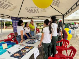 La fotografía muestra un evento comunitario al aire libre, organizado bajo una carpa con el rótulo “Alcaldía de Tame”. En el centro se observa una mesa con materiales informativos, folletos y carteles, acompañada por un letrero que dice “Fiscalía”, lo que indica la participación de esta institución en la actividad.Varias personas están reunidas alrededor de la mesa, interactuando y observando los recursos disponibles. A la izquierda se distinguen materiales coloridos con ilustraciones tipo caricatura y textos educativos, probablemente dirigidos a estudiantes o visitantes jóvenes. En el fondo aparecen otras carpas, sillas rojas y grupos de asistentes, lo que refuerza la idea de que se trata de una feria comunitaria o jornada de divulgación institucional.
