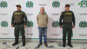 Se observa al capturado en el centro y de frente. A cada costado un funcionario de Policía Nacional. Atrás un pendón de Policía Nacional